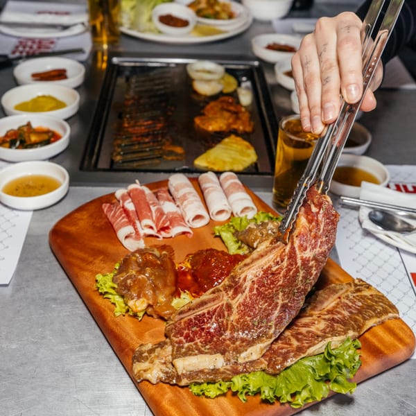 A hand uses tongs to place raw marinated Korean BBQ short ribs onto a hot grill.