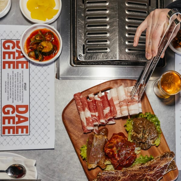 A hand uses tongs to place raw Korean BBQ meats onto a grill, with banchan and beer nearby.