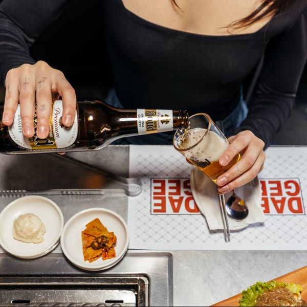 Hands pouring a Kloud Premium beer into a glass at a DAE GEE KOREAN BBQ table.